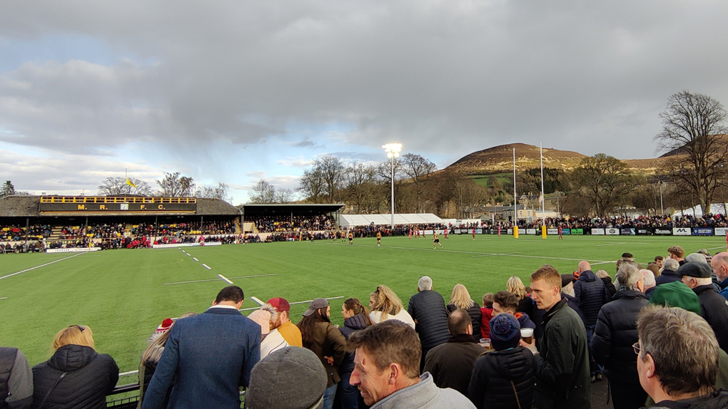 A crowd surrounds the Greenyards pitch, watching the Melrose Sevens rubgy tournament with the Eildon hills overlooking