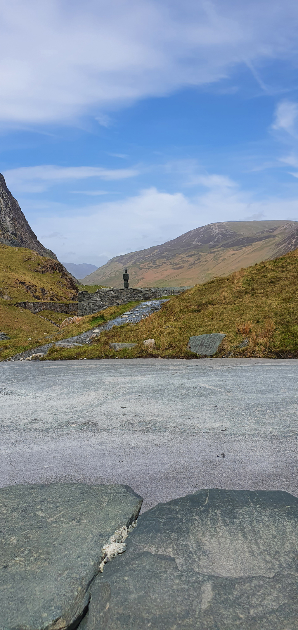 In the middle distance a slate statue sits on a slate wall, at the foot of a mountain. In the distance mountains