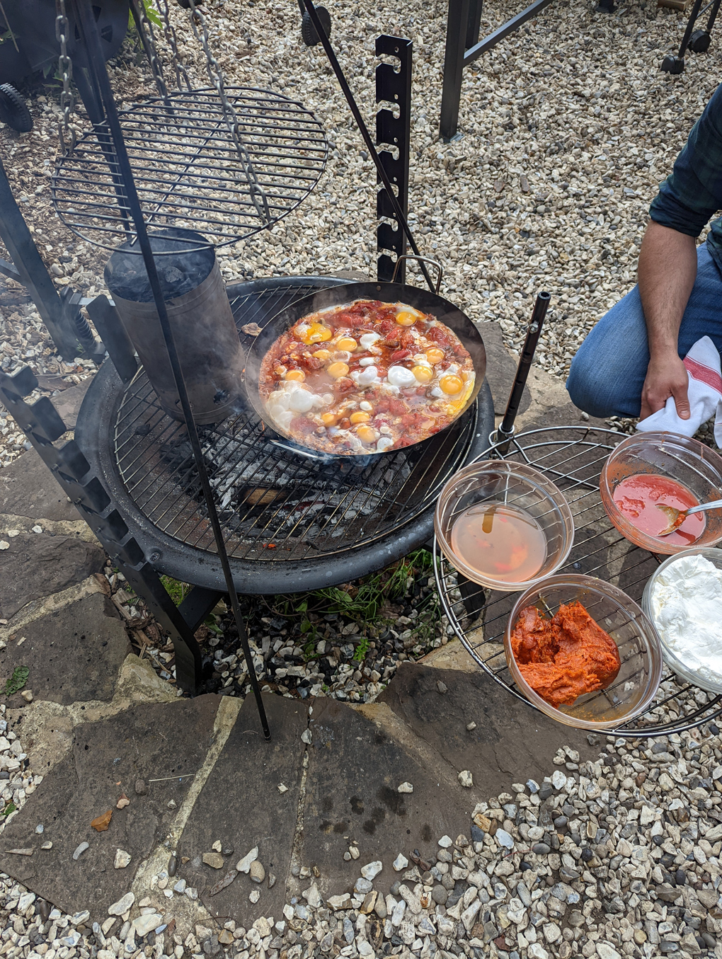 Shakshuka being cooked on a barbecue with a plate of additional ingredients next to it