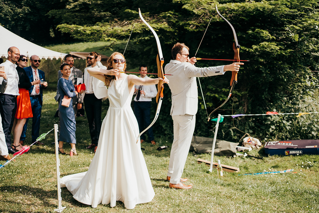 A bride and groom back to back holding bows at full draw with arrows pointed off screen