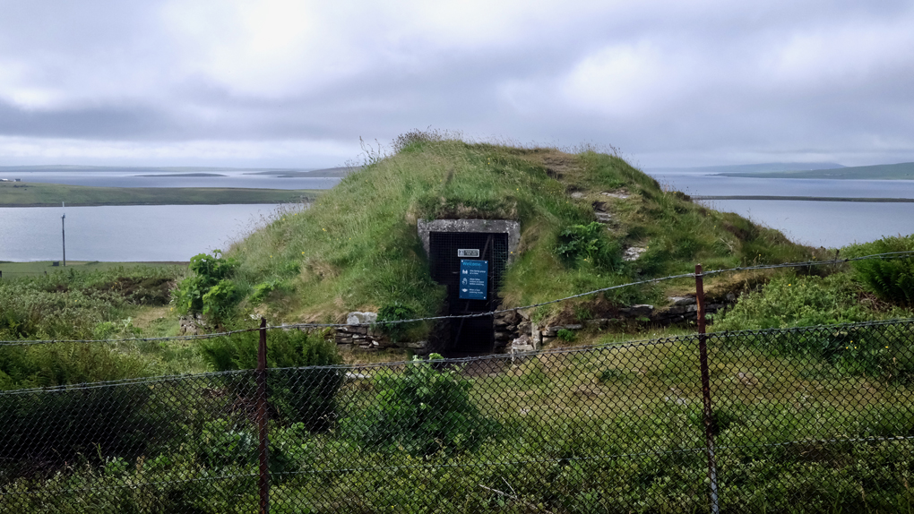 A small grassy cairn with an ancient stone entranceway, and a modern metal gate, stands high on a hillside overlooking the sea and small islands beyond.