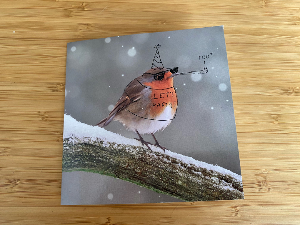 A greetings card with a photo of a robin in the snow. Someone has drawn on a party hat, sunglasses, a party horn and a t-shirt saying LET'S PARTY!