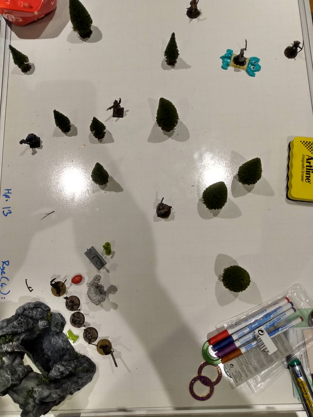 The whiteboard we use for D&D with a cave mouth and miniature trees scattered over it. Several miniatures line up for combat.