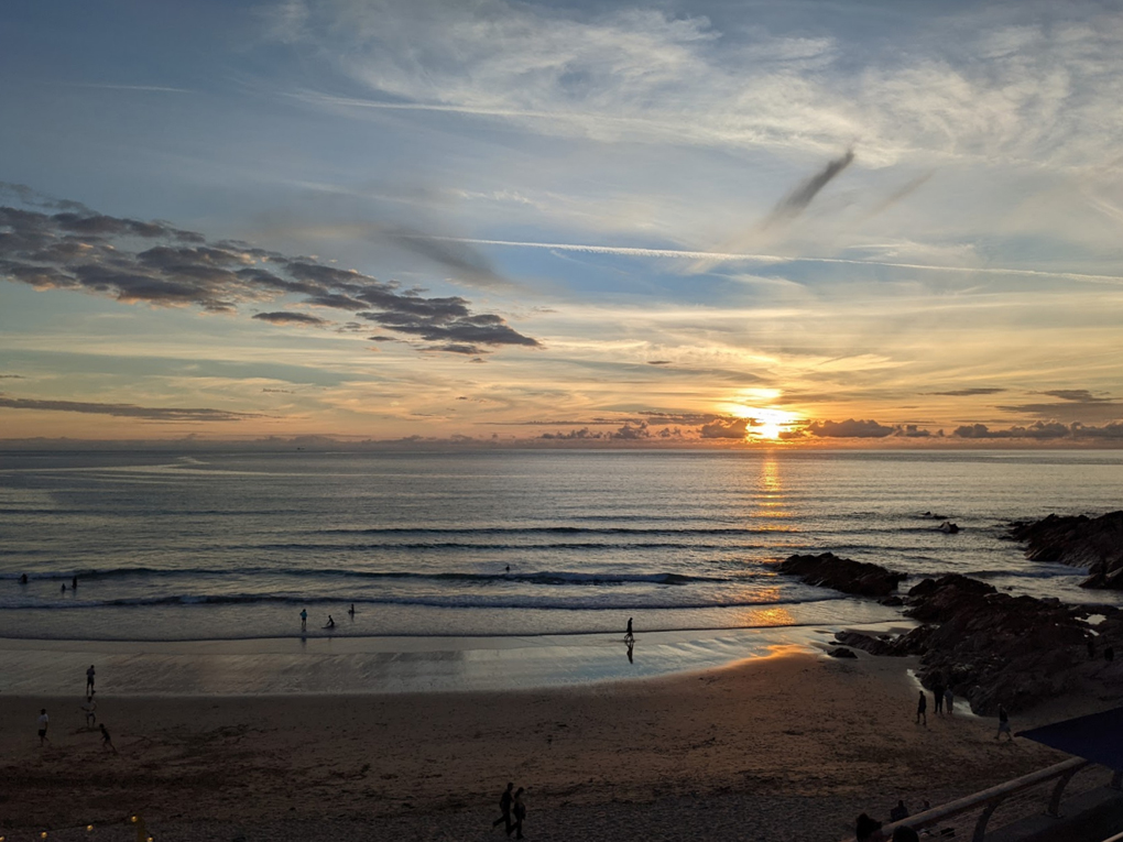 A bright orange sun sets amongst whispy clouds above a calm blue-grey sea