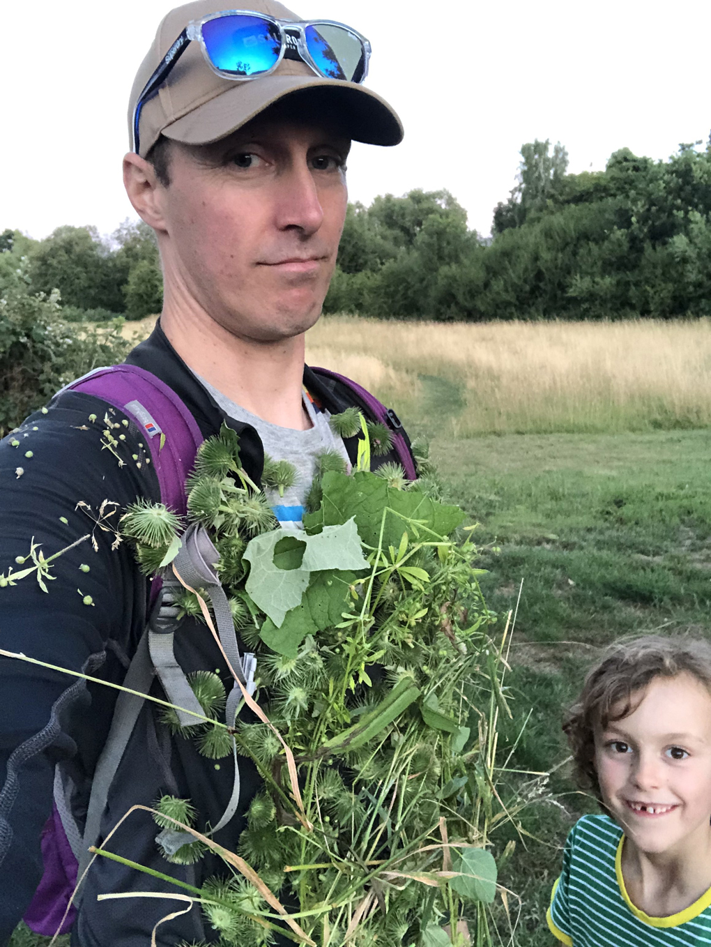 An unimpressed man in a field, with a large amount of sticky foliage attached to his chest and front, with a small child with blond hair in the corner, with a mischievous smile on his face.