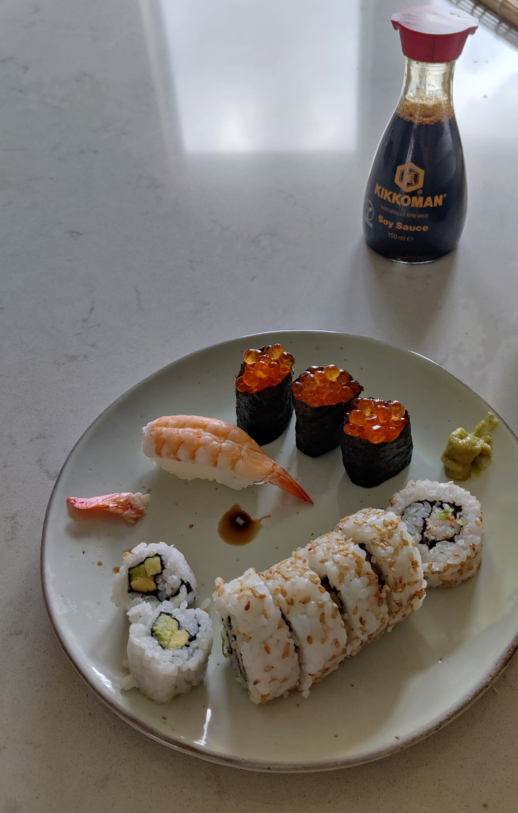 A plate of homemade sushi on a white stone kitchen counter, including salmon roe gunkan, ebi prawn nigiri and crab rolls.