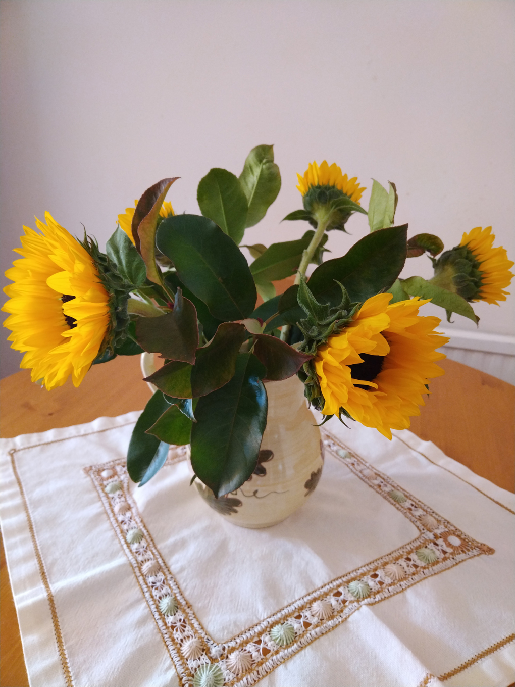 A jug with sunflowers and dark green foliage on a tablege