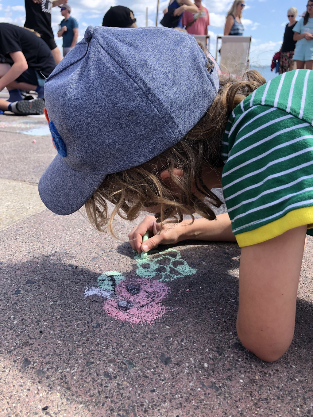 A blonde boy wearing a blue cap and a green and white striped t-shirt using chalk to draw fruit and vegetables on the pavement.