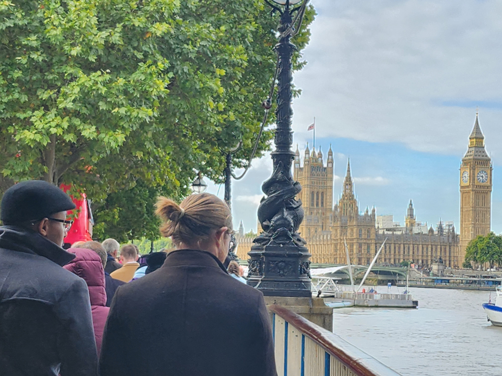 On the left, the Palace of Westminster sits across the river, the flag flies at half mast. The right side of the photo is a queue of people, two abreast as far as can be seen