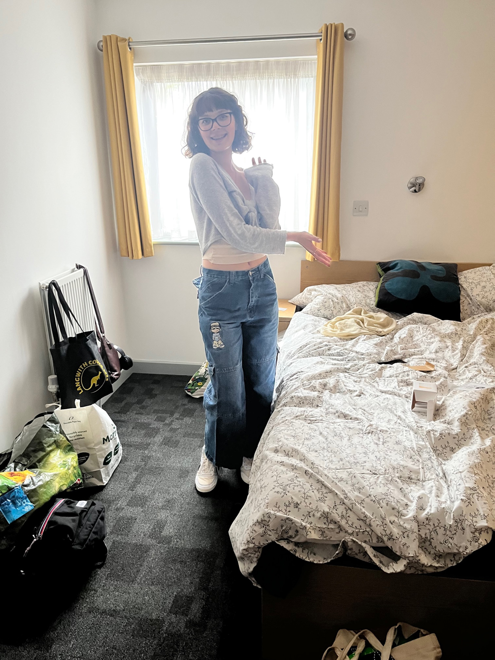 A happy looking young women in jeans and a grey cardigan points at an untidy bed