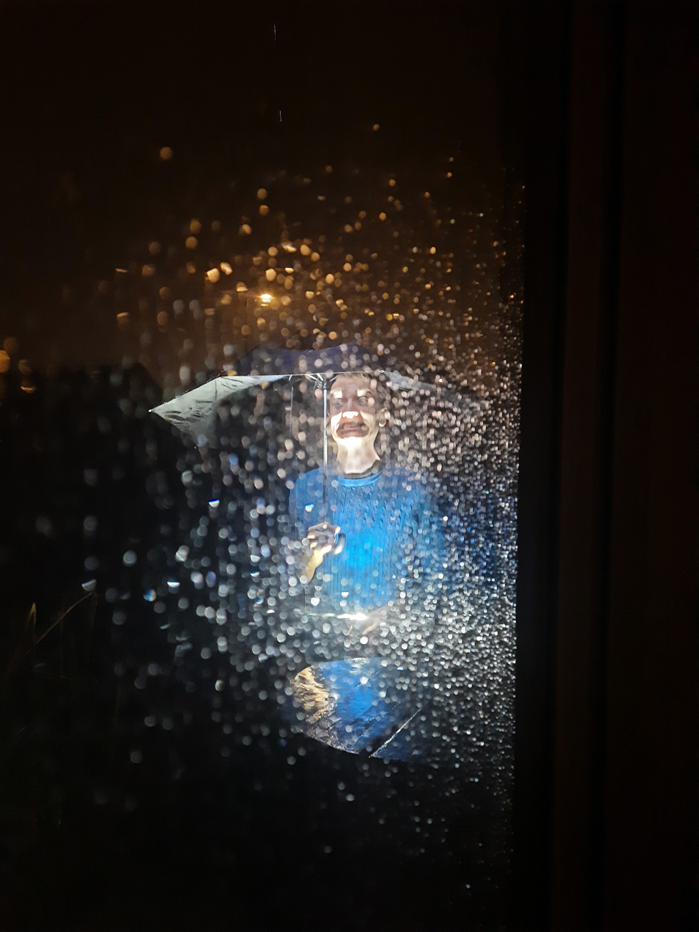 A photo taken through a window at night. The rain drops on the window and falling obscure a man under an umbrella. A torch illuminates parts of his face and the water droplets creating an uncanny effect.