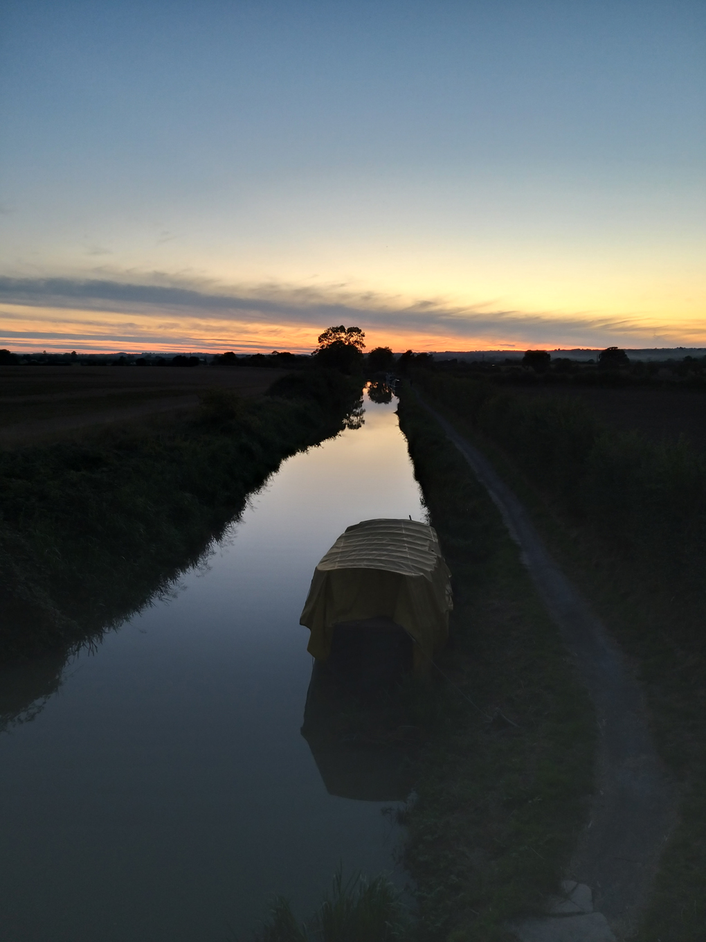 Sunset afterglow on the horizon, canal stretching away into the distance with a couple of trees and a solitary boat reflecting into it.