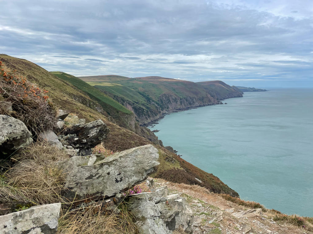 Rocks in the foreground with a view behind of grassy cliffs and the sea. Clouds in the sky