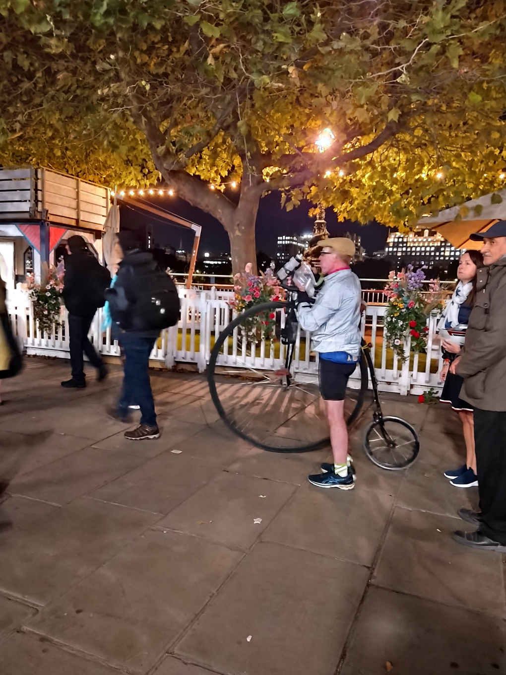 A man eating a boiled from a plastic bag and pushing a modern Penny Farthing