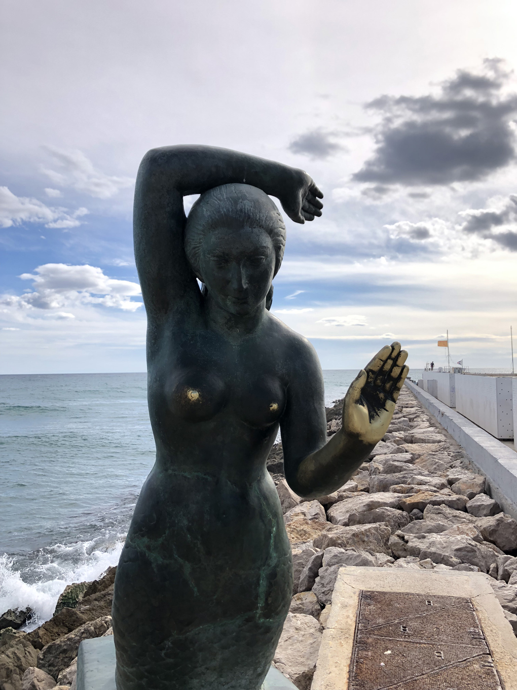 A bronze statue of a mermaid against the backdrop of the Mediterranean, with her left palm held forward.