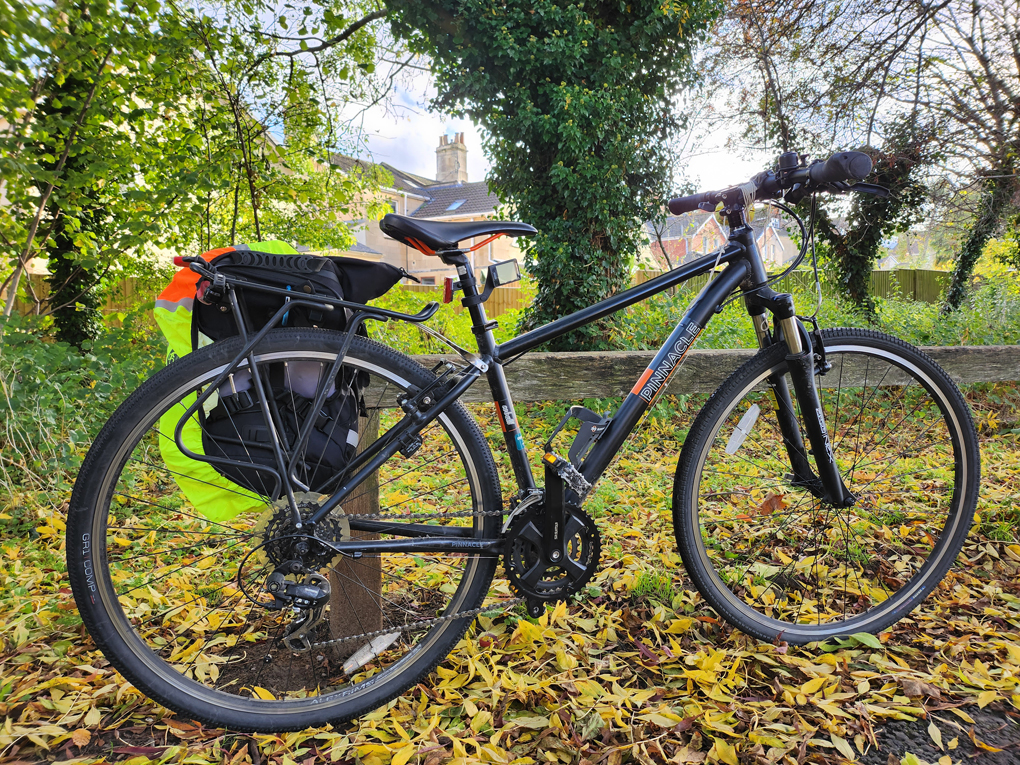 A mostly black bike leans against a wooden bench. Yellow and orange leaves lie on the path.