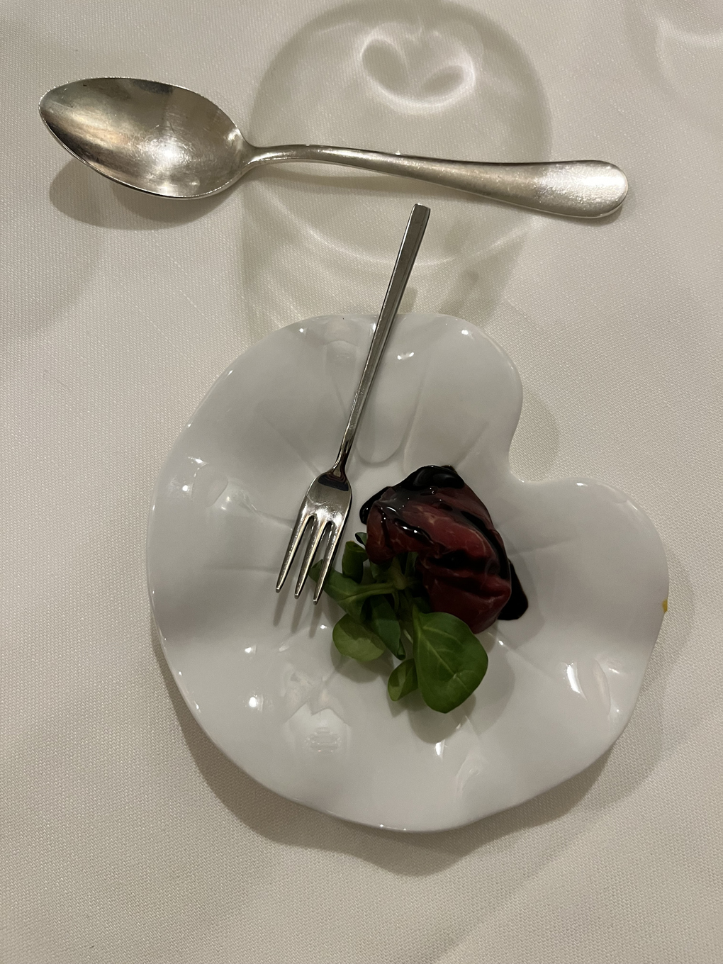 A bite-sized piece of smoked meat on a clean white plate. There is polished silver cutlery next to the plate.
