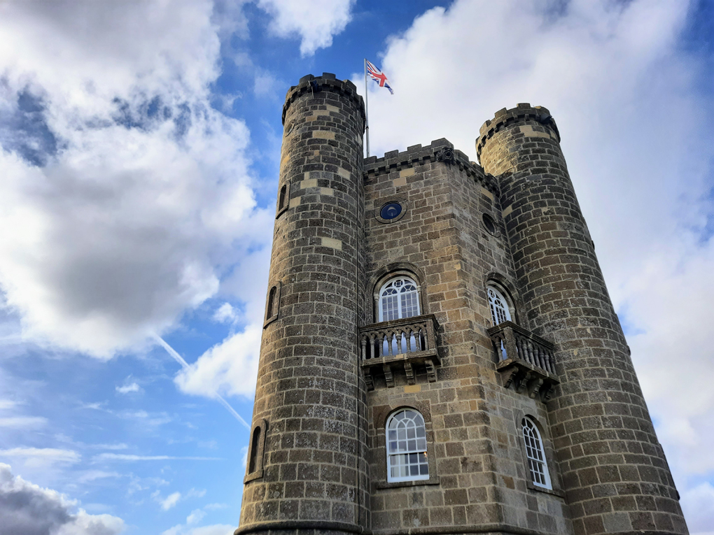 Looking up at an old stone tower with small turrets on either side. A Union Jack flag flies at the top against a backdrop of blue sky and fluffy clouds