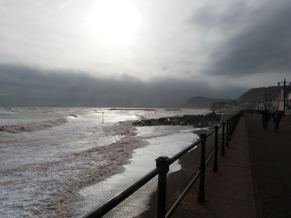 Waves breaking along the Sidmouth seafront, with dark clouds covering the sun and looming above the western horizon