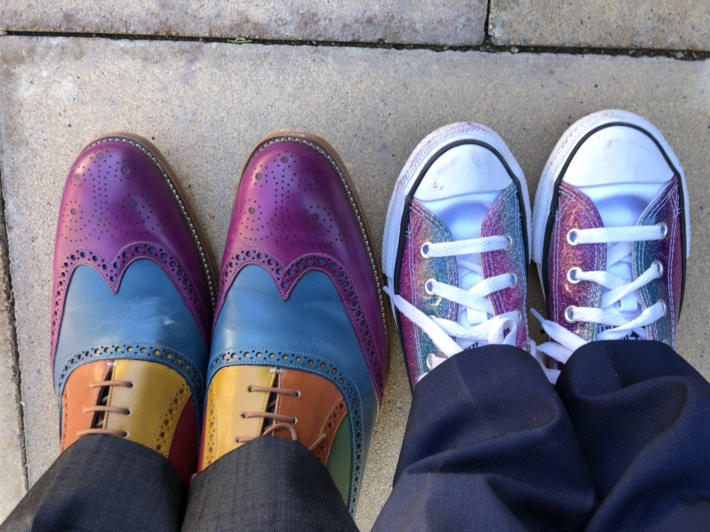 Two pairs of feet, viewed from above, wearing colourful shoes; an adult wearing patent leather brogues with each panel a different colour, and a child wearing Vans with sparkly purple and blue panels.