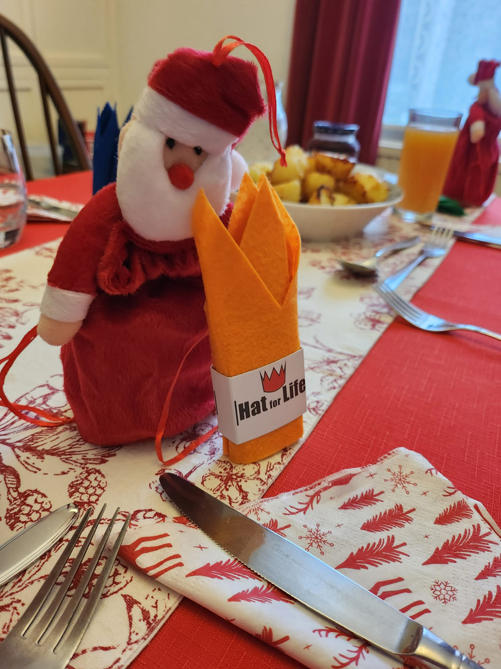 A small santa figure made of fabric sits besides a cloth hat for life Christmas crown. The figure and crown sit on a dining table.