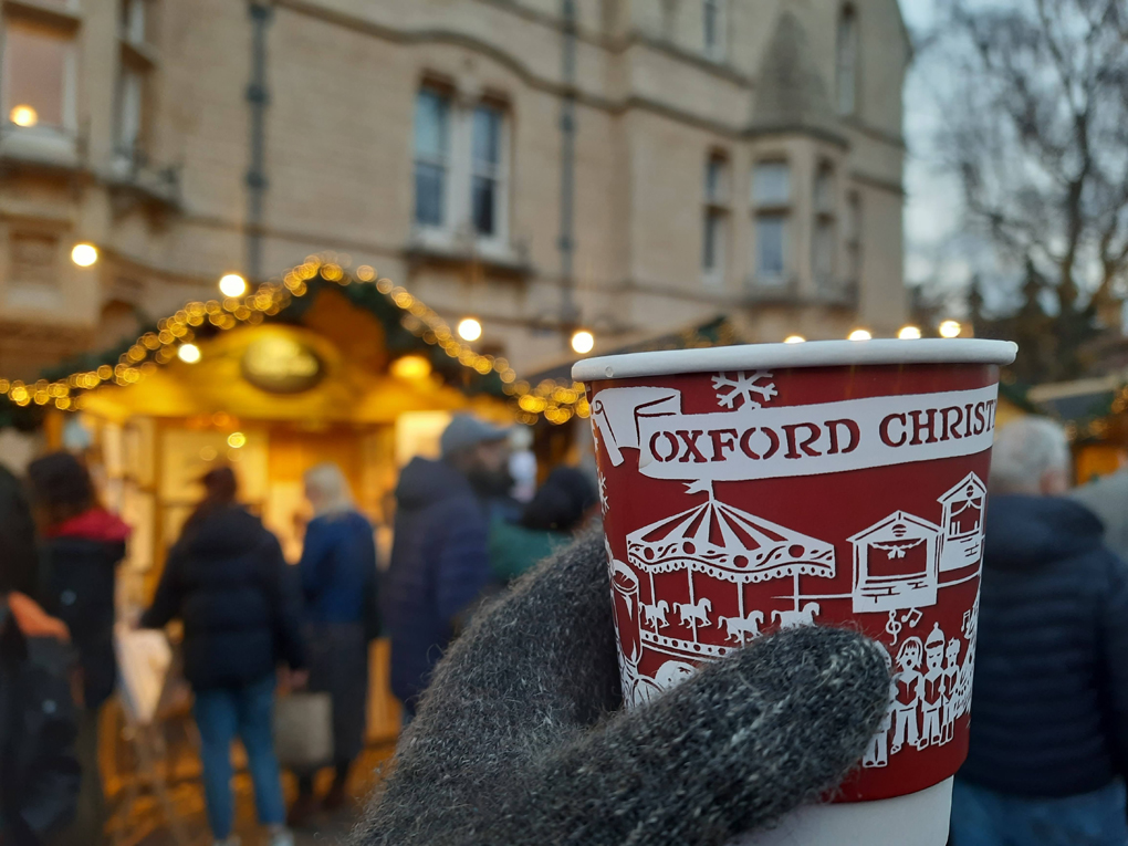 A gloved hand holding a papercup of warm drink with the phrase 'Oxford market' on the side.