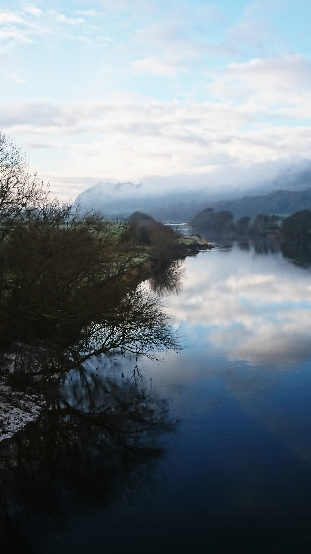 A lovely sleepy river on a very cold and frosty day, reflecting the clouds in a soft blue sky, the distant hills striped with a blanket of fog.