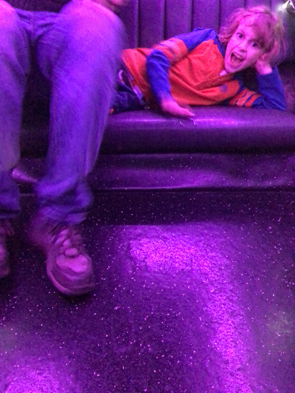 A boy lying on a sofa yelling as someone who is mostly out of shot sits down on his legs.