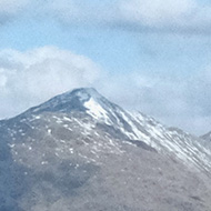 A small loch high in the grassy hills, with snow covered mountains beyond.