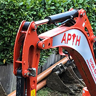 A muddy patch, with foundation trenches dug for a new extension, a mini-digger pulling up the mud and rubble, with a high wooden fence and green hedge in the background.