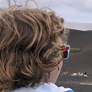 A muzzled camel and a small boy with long dark blonde hair looking at each other, with a desolate  volcanic vista in the background.