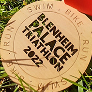 A wooden medal etched with Blenheim Palace Triathlon 2022 with a red ribbon woven around it.