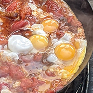 Shakshuka being cooked on a barbecue with a plate of additional ingredients next to it