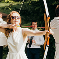 A bride and groom back to back holding bows at full draw with arrows pointed off screen