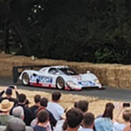 A field of people watching a low white sports car with blue and red livery travelling at speed 