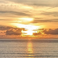 A bright orange sun sets amongst whispy clouds above a calm blue-grey sea