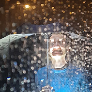 A photo taken through a window at night. The rain drops on the window and falling obscure a man under an umbrella. A torch illuminates parts of his face and the water droplets creating an uncanny effect.