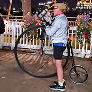 A man eating a boiled from a plastic bag and pushing a modern Penny Farthing