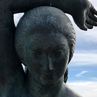 A bronze statue of a mermaid against the backdrop of the Mediterranean, with her left palm held forward.