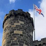 Looking up at an old stone tower with small turrets on either side. A Union Jack flag flies at the top against a backdrop of blue sky and fluffy clouds
