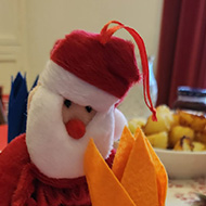 A small santa figure made of fabric sits besides a cloth hat for life Christmas crown. The figure and crown sit on a dining table.