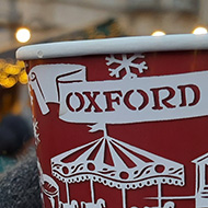 A gloved hand holding a papercup of warm drink with the phrase 'Oxford market' on the side.