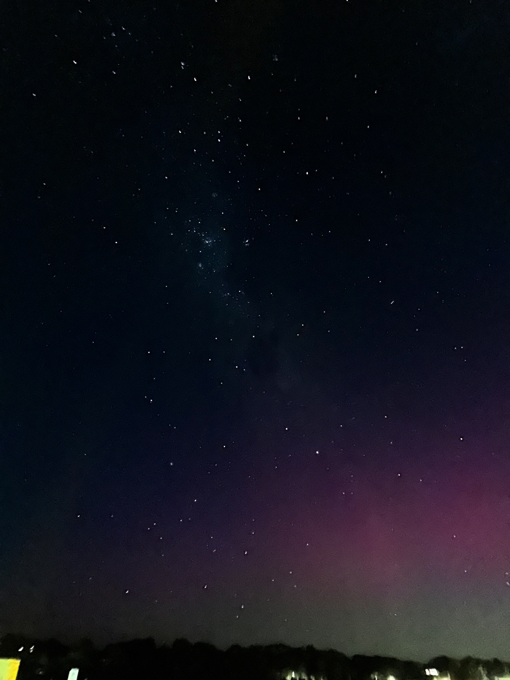 Stars clustering in a band vertically. The Milky Way. Bottom right is a red and green haze, the Aurora Australis. Some building in the distance with lights on at the bottom.