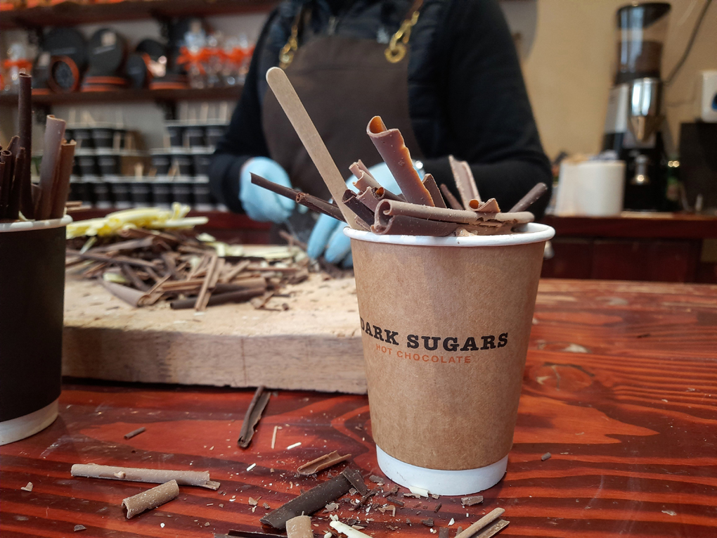 A cup of hot chocolate piled high with white milk, and dark chocolate shavings. In the background is a another large pile of chocolate.