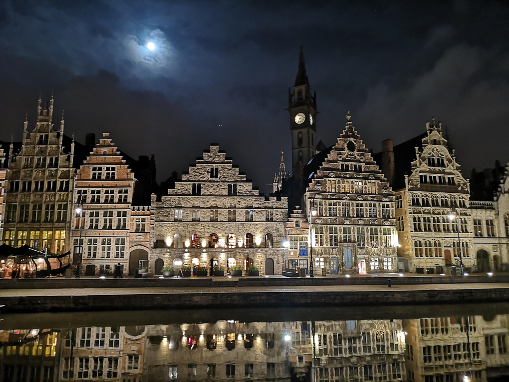 Night view of buildings in Ghent reflected in a canal with the moon behind.