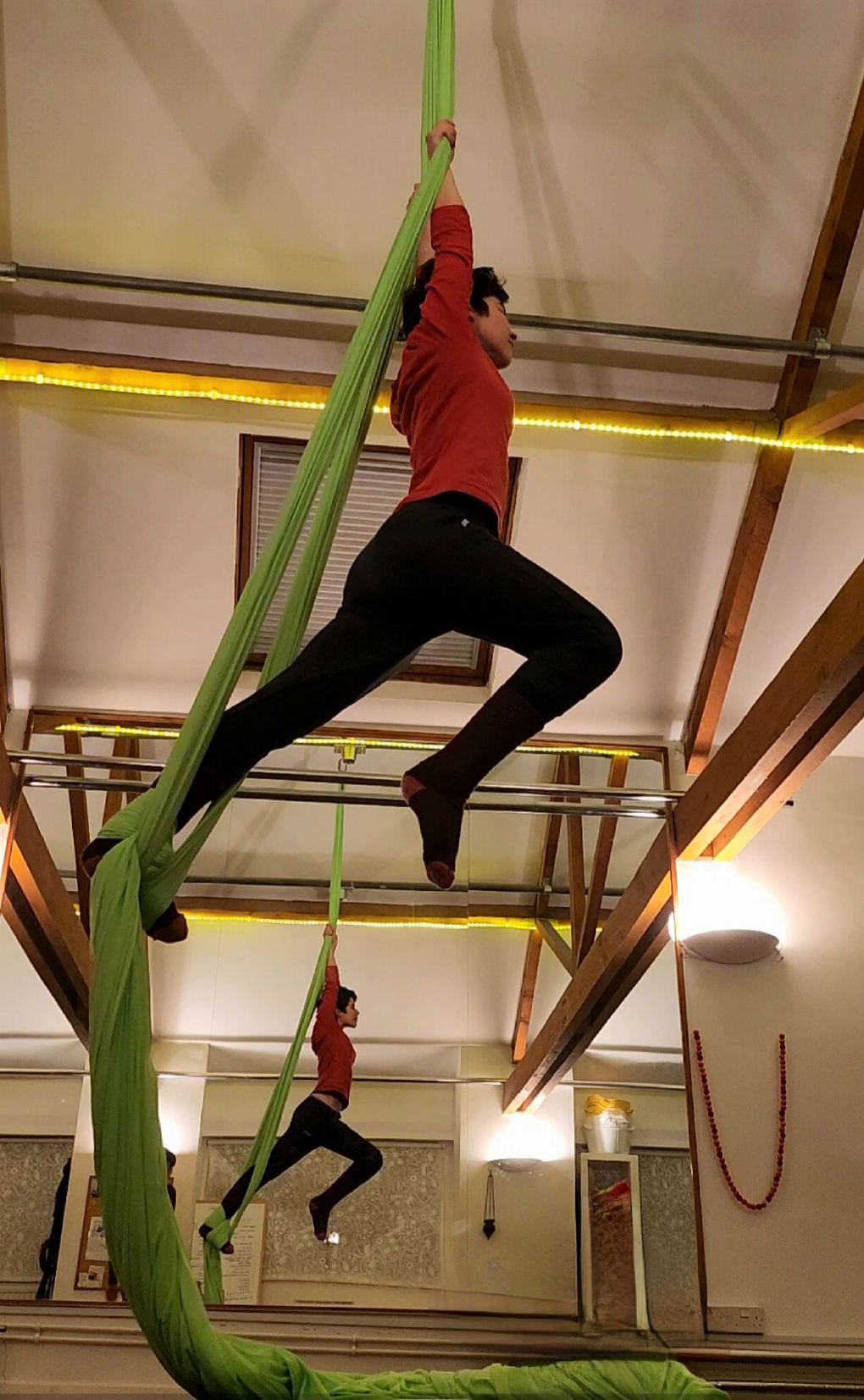 A short haired female holds onto a green silk hanging from the ceiling, one foot is wrapped up by the silk, the other leg is held forward in an attempt to be elegant. A mirror shows her reflection in the background