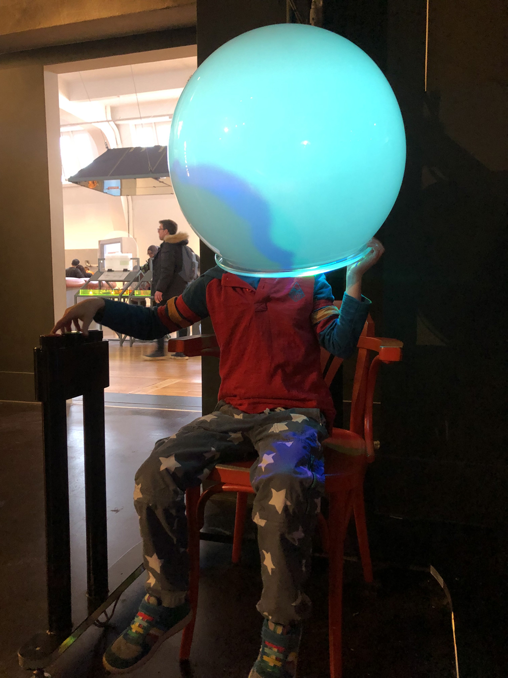 A young boy sitting on a chair in the Science Museum, with a big, opaque spherical helment over his head, emitting a soft blue glow.