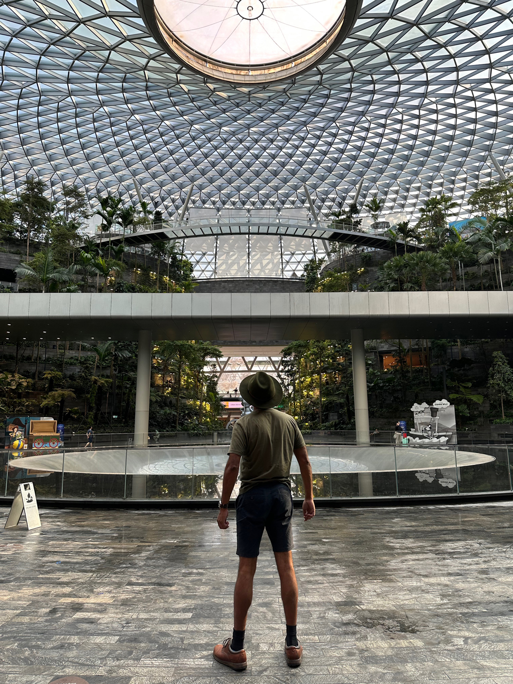 A man in an olive coloured t-shirt and blue shorts with a green felt hat photographed from behind. The inside of a domed structure ahead with an inactive water fountain.