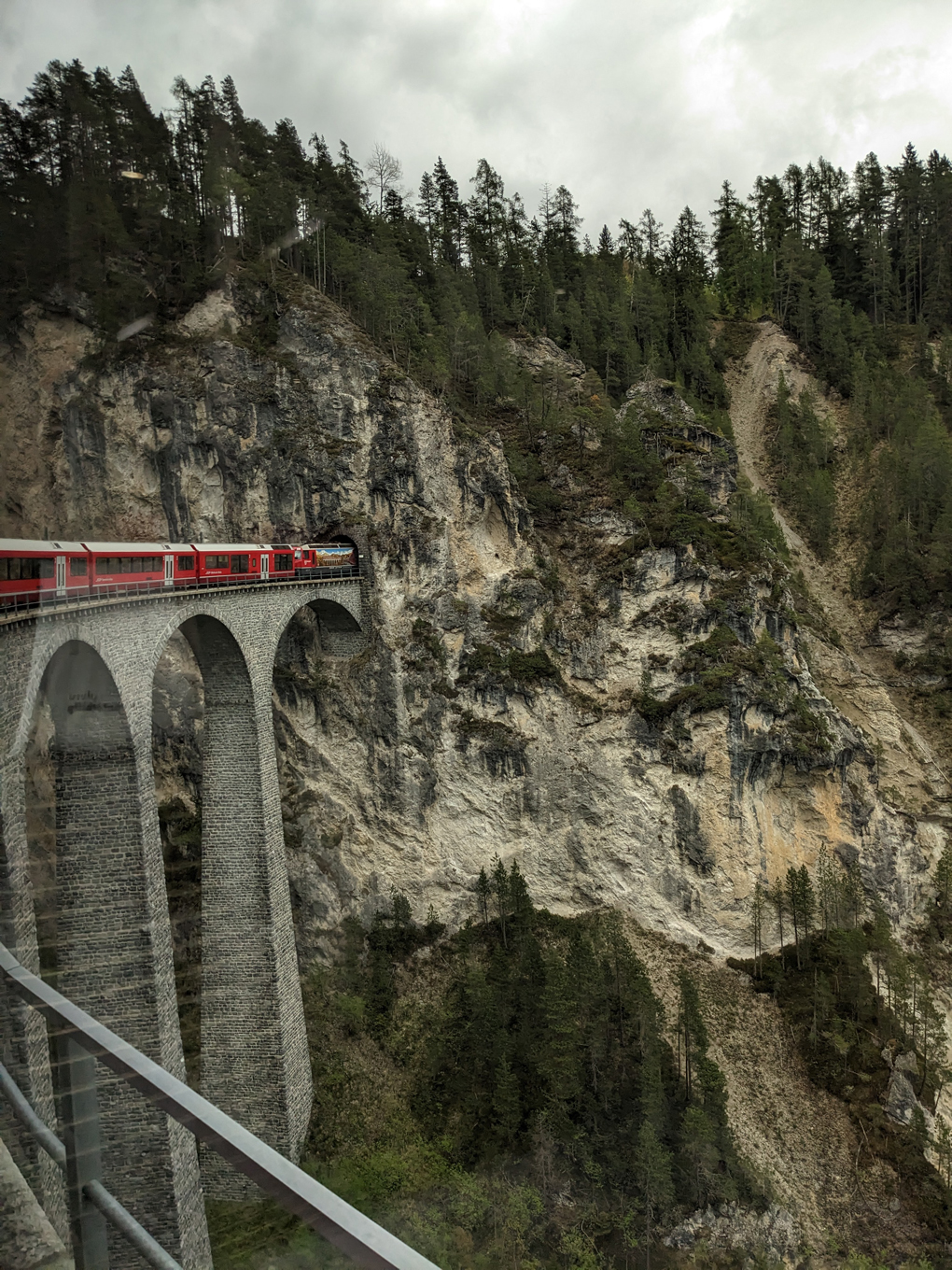 The front of a train going over a viaduct into a tunnel through a mountain in the alps. Taken from the back of the train.