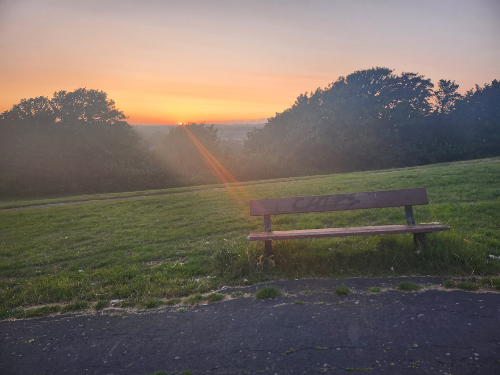 A thin orange, yellow, red sun beam strikes a park bench. A weak yellow beam is fading as the sun has nearly disappeared beyond the horizon. The horizon consists of a row of rooftops and trees in the distance